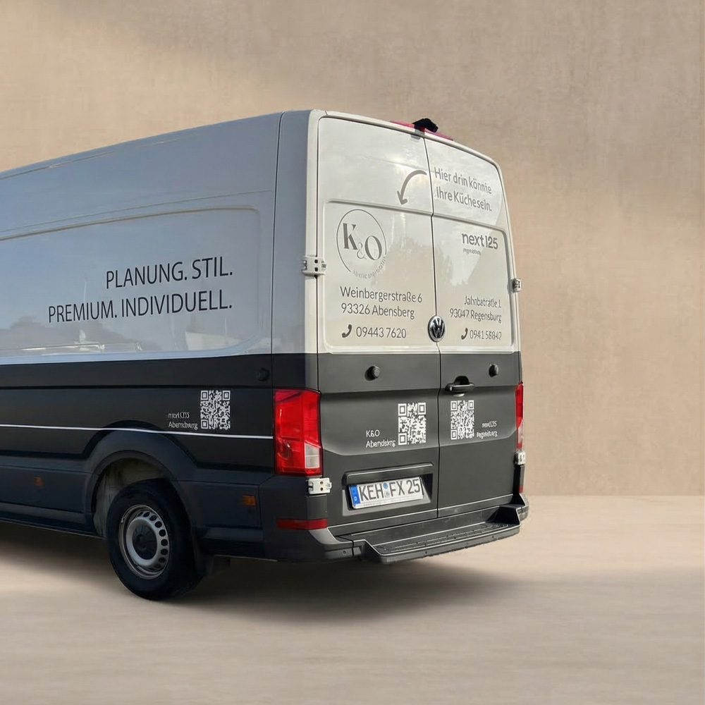 Heckansicht eines VW Transporters mit einer Firmen-Beklebung. Die komplette untere Hälfte des Transporters ist in einer schwarz-matten Folie foliert mit 3 verschiedenen QR-Codes. Die obere Hälfte ist weiß und mit dem Logo, Adresse und Telefonnummer des Unternehmens 