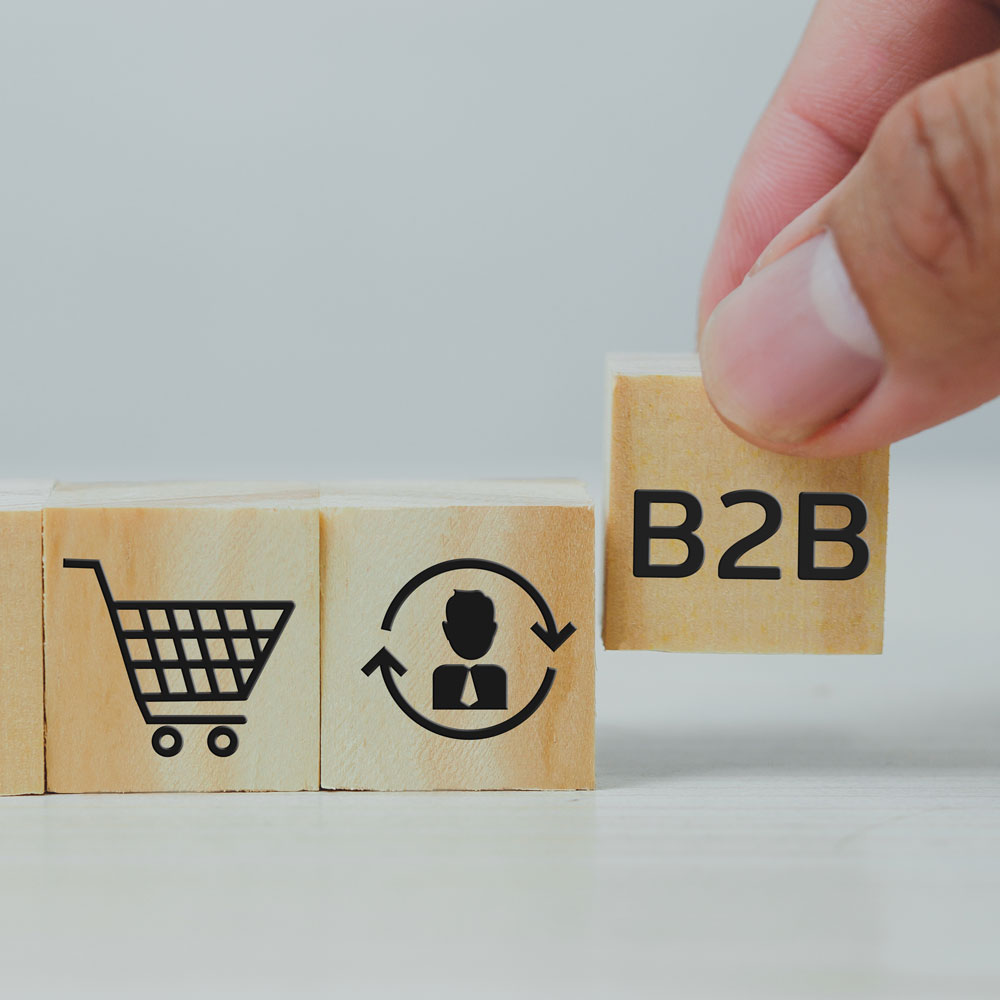 Drei Holzwürfel mit unterschiedlichen schwarzen Icons stehen in einer Reihe auf einer weißen Oberfläche. Der linke Holzwürfel mit einem "Einkaufswagen"-Icon, in der Mitte ein Icon mit einer Person und Pfeile, die sich um die Person drehen. Der rechte Würfel wird gerade von zwei Fingern angehoben und schwebt etwas über den anderen, hat eine "B2B" Aufschrift.